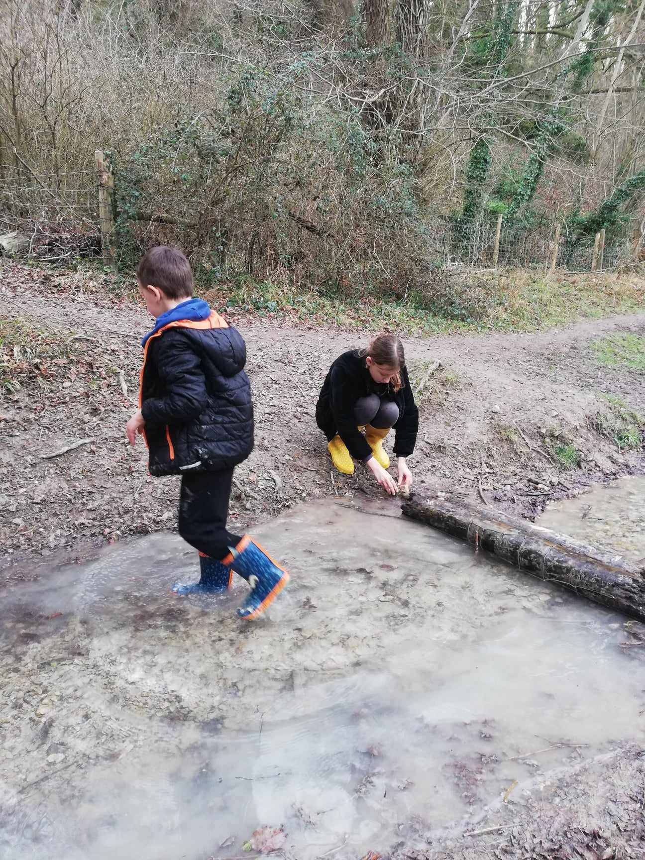 Dwoje dzieci bawi się w błotnistym strumieniu w lesie; chłopiec w niebieskich kaloszach z pomarańczowymi akcentami wchodzi do wody, dziewczynka w żółtych kaloszach kuca obok powalonego pnia.