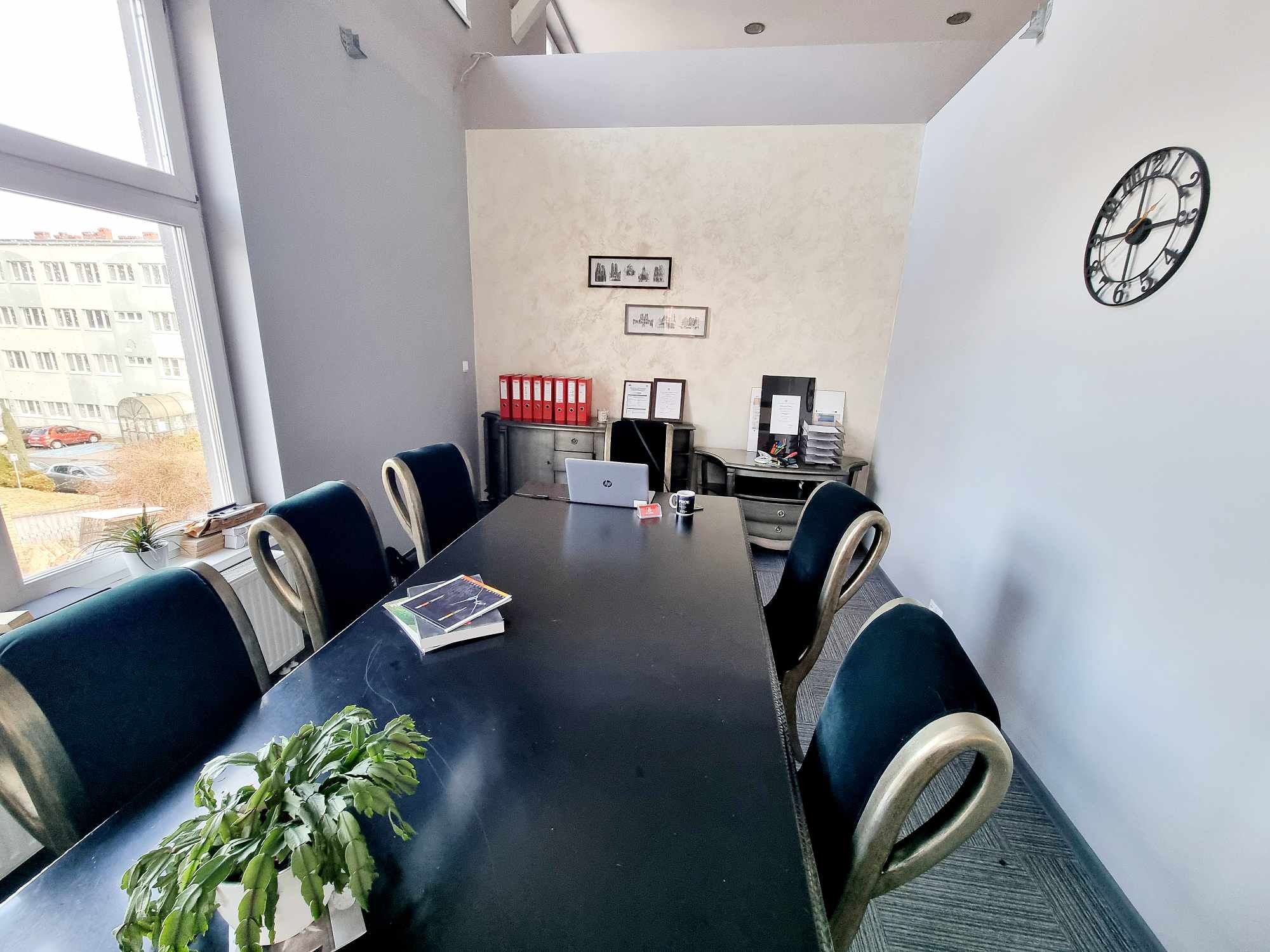 Sala konferencyjna z dużym, czarnym stołem, zielonymi krzesłami i rośliną doniczkową na stole. W tle biurko z czerwonymi segregatorami, laptopem i dekoracjami ściennymi.