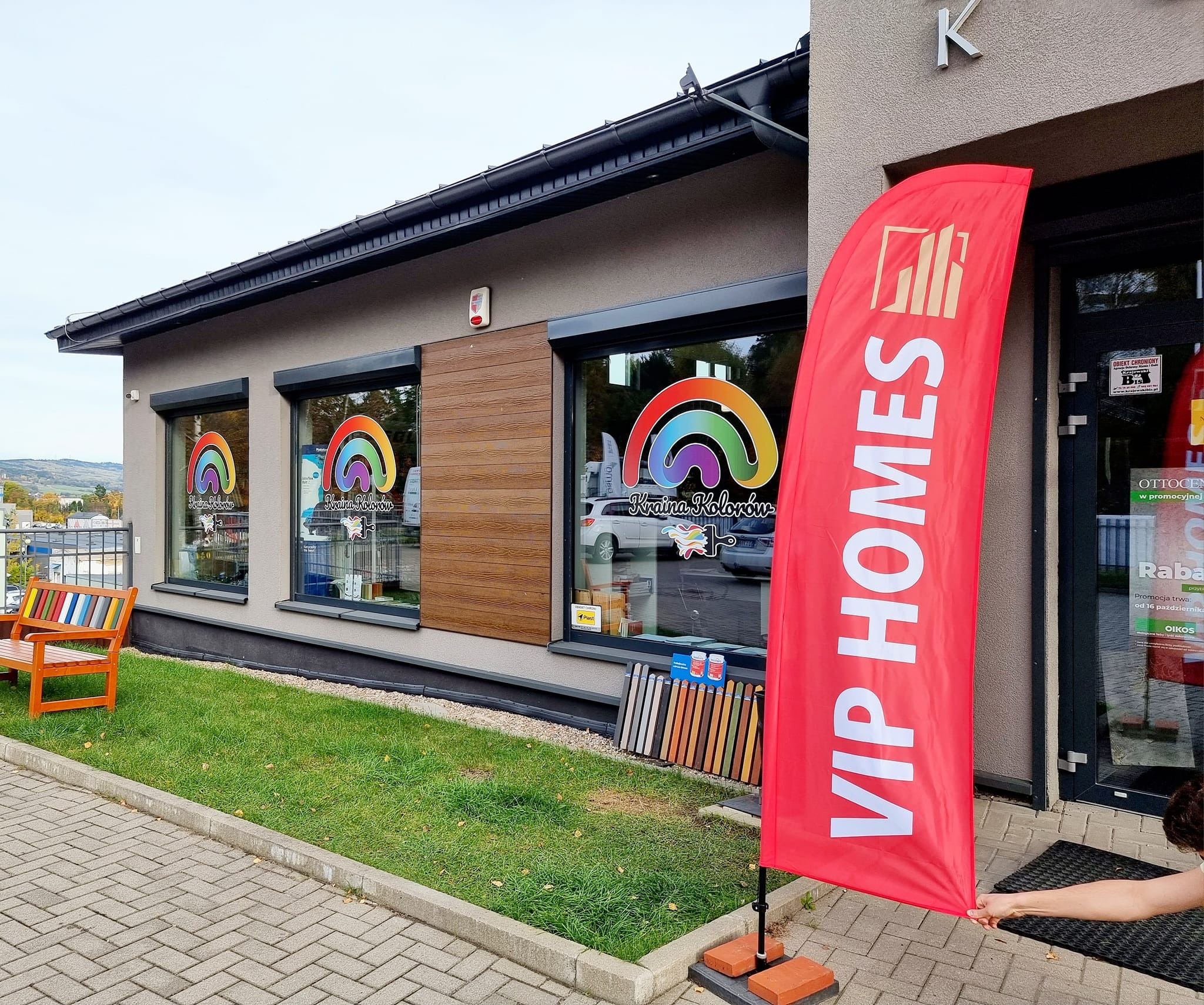 Elewacja budynku firmy 'Kraina Kolorów' z logo tęczy w oknach, czerwona flaga 'VIP HOMES' i próbki kolorów na trawniku.