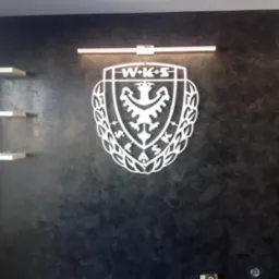 Ściana z efektem dekoracyjnym w odcieniach szarości, podświetlona listwą LED, z zamontowanym białym logo WKS i trzema półkami po lewej stronie.