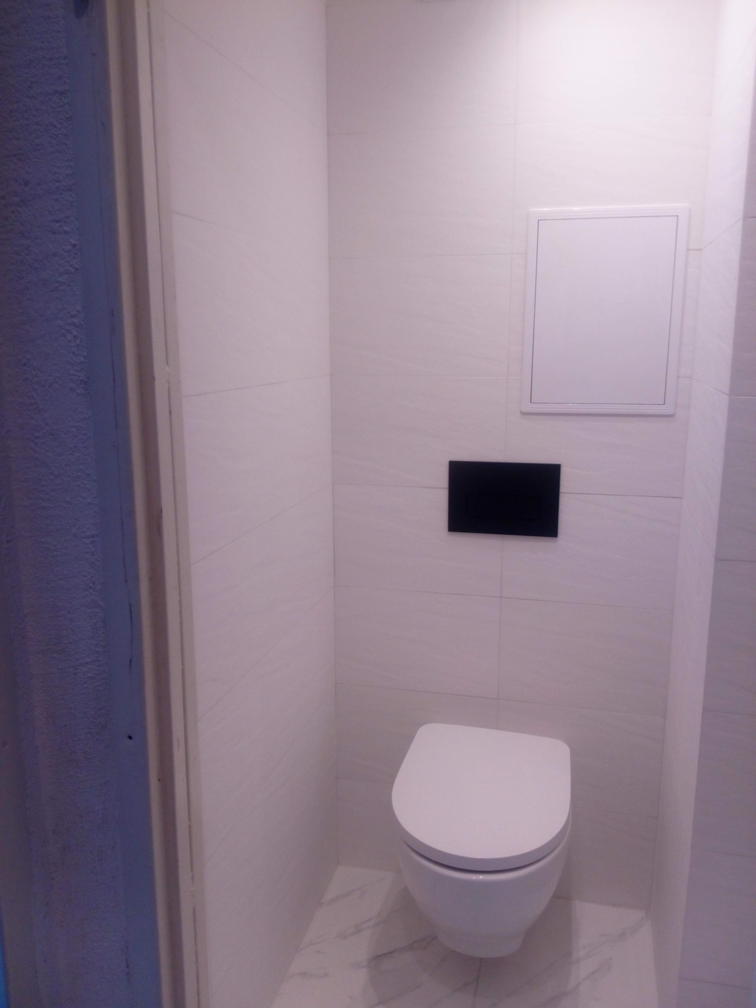 Nowoczesna toaleta podwieszana z czarnym przyciskiem spłukującym i białymi płytkami imitującymi marmur, widoczny biały właz rewizyjny.