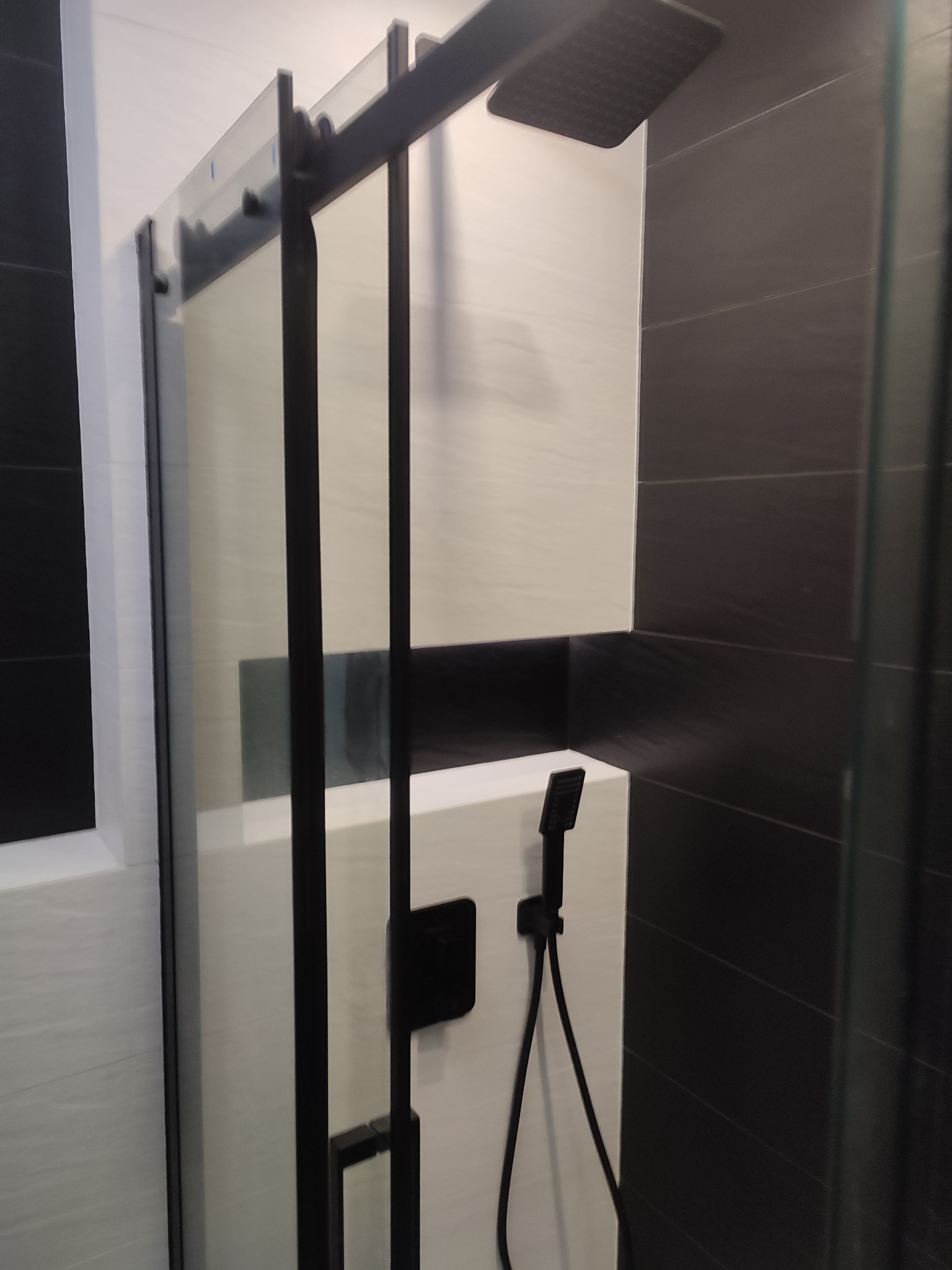 Nowoczesna kabina prysznicowa z czarnymi profilami i deszczownicą, biało-czarne płytki w tle.