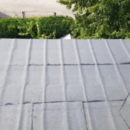 Widok z góry na szary, płaski dach pokryty papą, z widocznymi łączeniami i niewielkimi uszkodzeniami, otoczony zielenią drzew i krzewów oraz elementami otoczenia budynku.