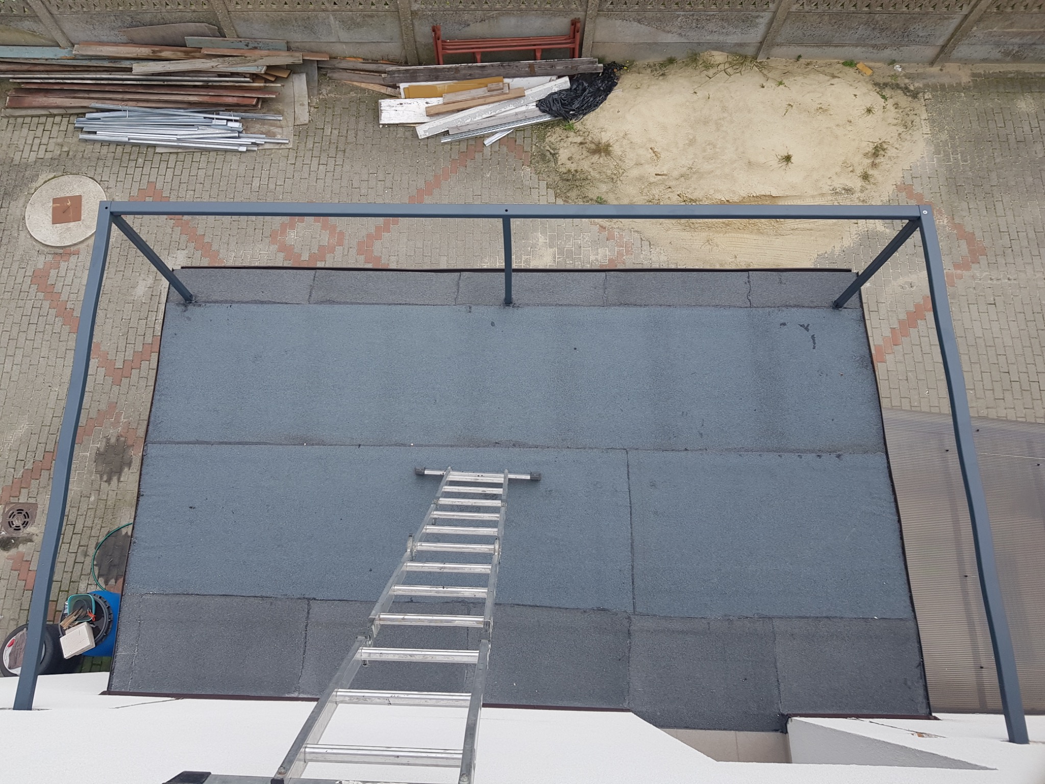 Widok z góry na płaski dach pokryty papą, z metalową konstrukcją zadaszenia i drabiną opartą o krawędź dachu. W tle widoczne materiały budowlane i teren z piaskiem.