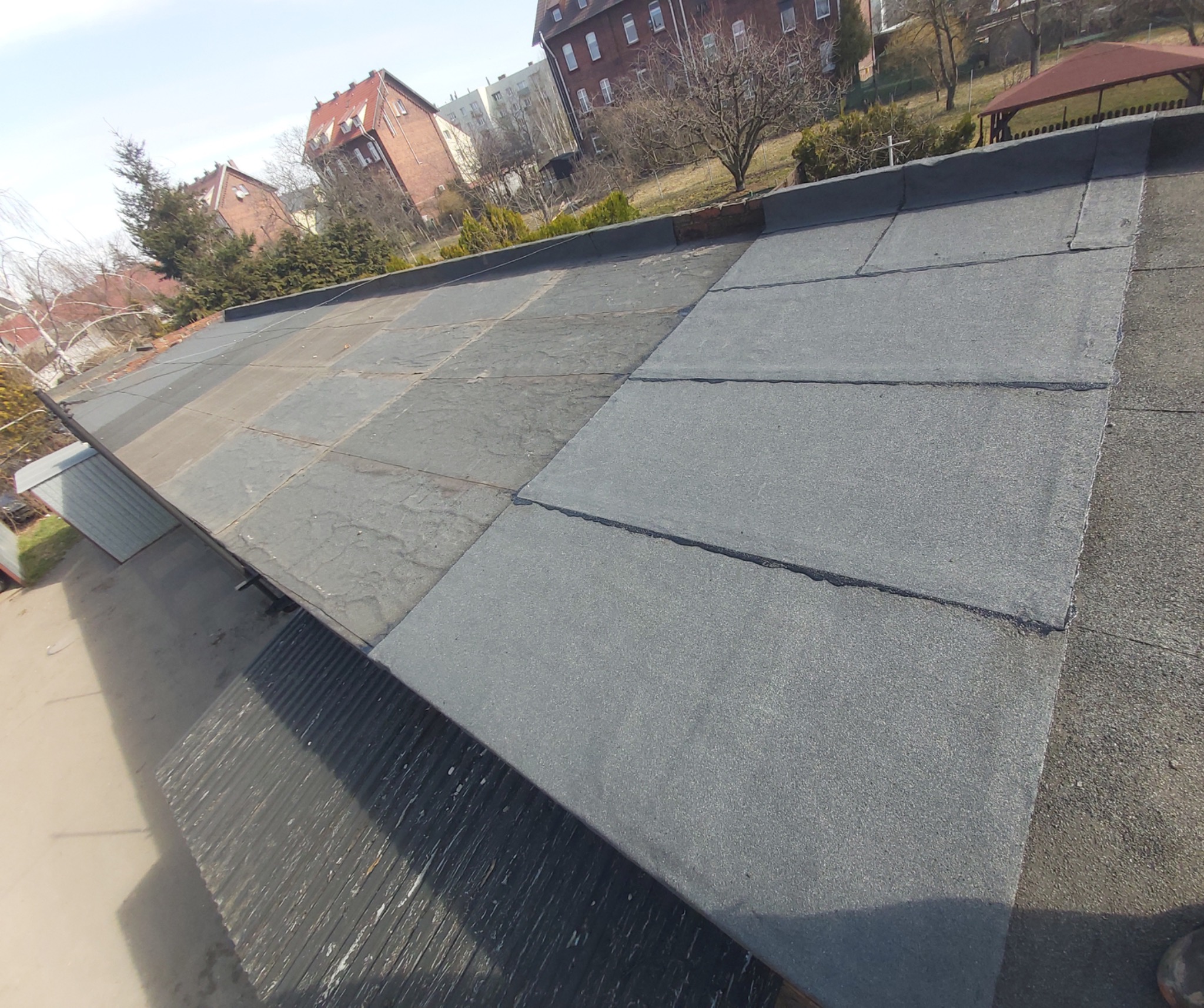 Płaski dach pokryty papą termozgrzewalną, widoczne połączenia między pasami papy, w tle budynki mieszkalne i drzewa.