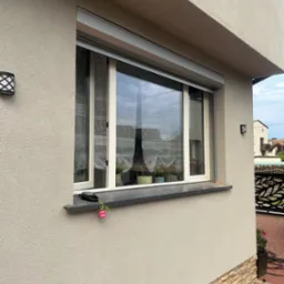 Zewnętrzny widok okna z roletą antywłamaniową, na parapecie leży zabawka w kształcie różowej świnki, elewacja domu w odcieniach beżu i szarości.