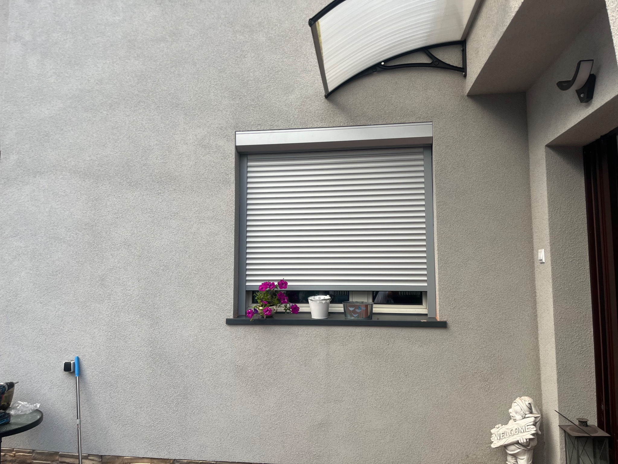 Szara fasada domu z zamkniętą roletą zewnętrzną, na parapecie kwiaty w doniczce, nad oknem daszek, obok drzwi wejściowe i dekoracyjny gipsowy krasnal z napisem Welcome.