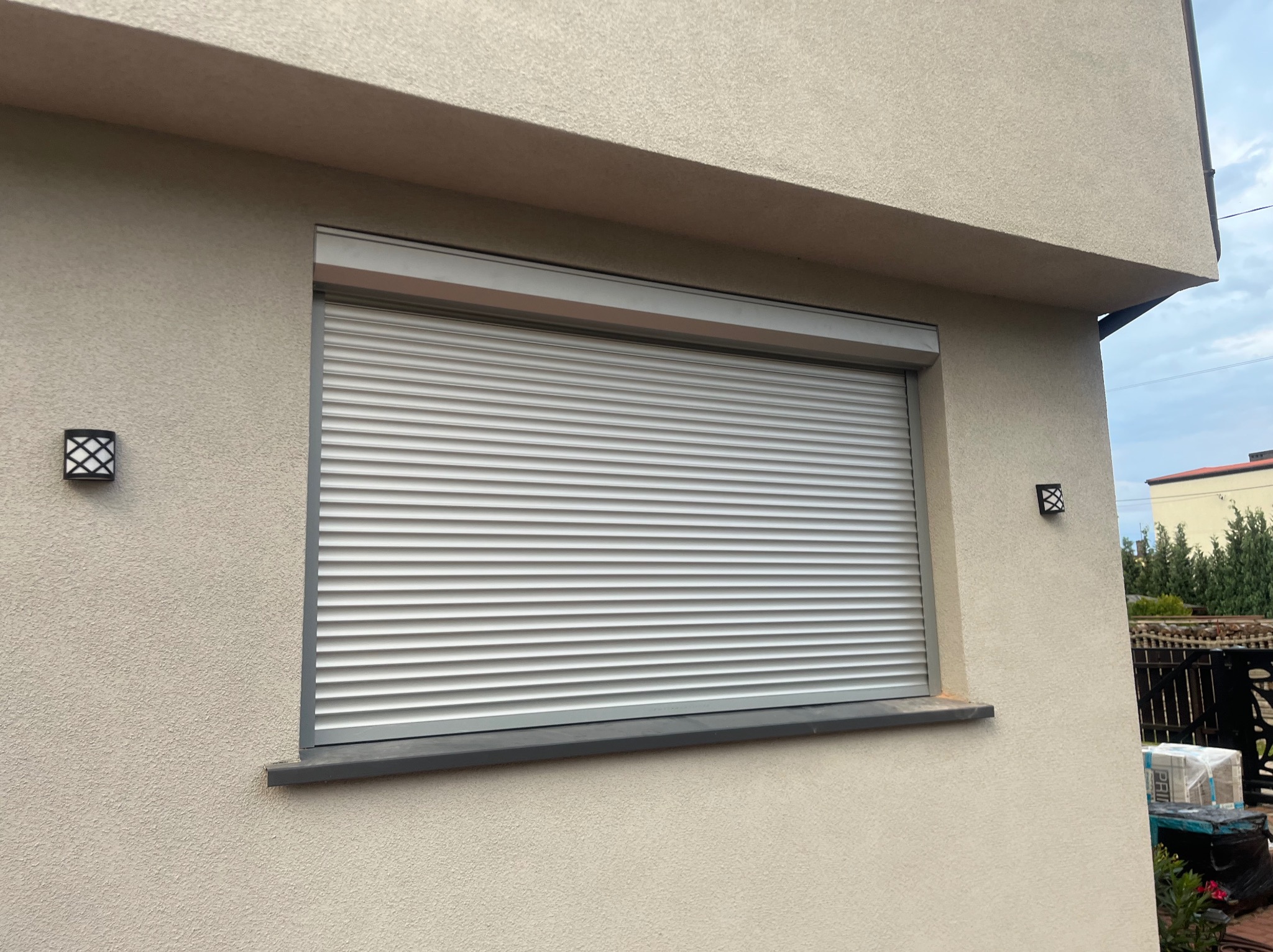 Zamknięta, szara roleta zewnętrzna na oknie budynku z beżową elewacją, widoczne lampy zewnętrzne po bokach okna.