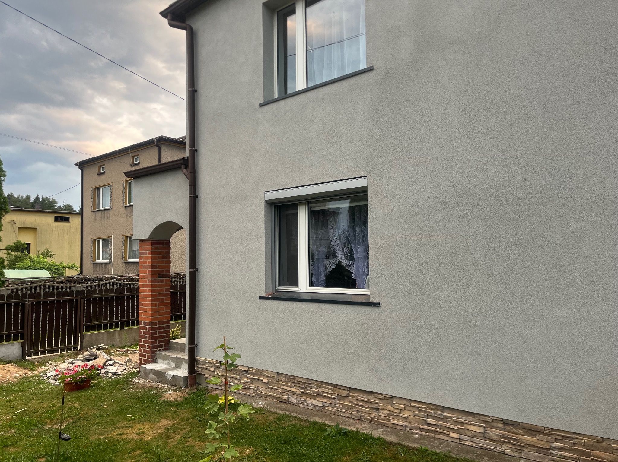 Szara elewacja domu jednorodzinnego w Siemianowicach Śląskich z częściowo opuszczoną roletą antywłamaniową na oknie parteru i widoczną dekoracyjną podmurówką z kamienia.
