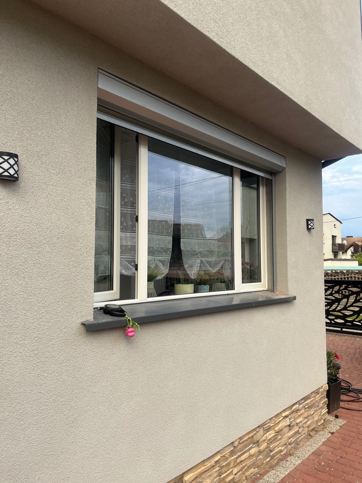 Zewnętrzny widok okna z roletą antywłamaniową, na parapecie leży zabawka w kształcie różowej świnki, elewacja domu w odcieniach beżu i szarości.