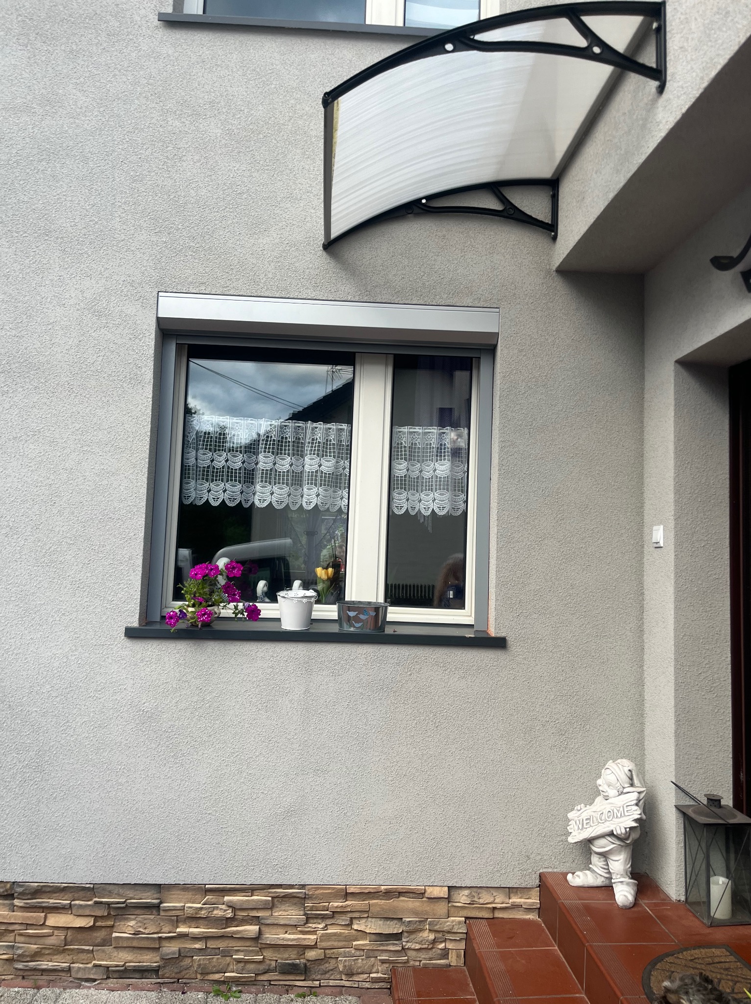 Szara fasada domu z oknem z częściowo opuszczoną roletą zewnętrzną, kwiatami w doniczkach na parapecie, dekoracyjną markizą nad wejściem i figurką krasnala z napisem WELCOME na schodach.