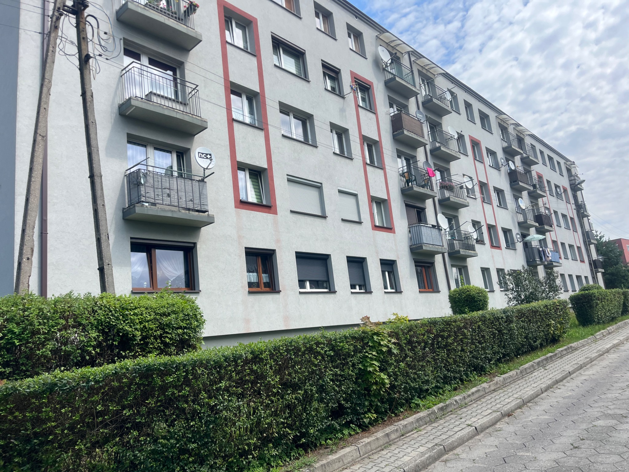 Wielopiętrowy budynek mieszkalny z balkonami, częściowo z roletami zewnętrznymi, widziany z poziomu ulicy, z przodu żywopłot.