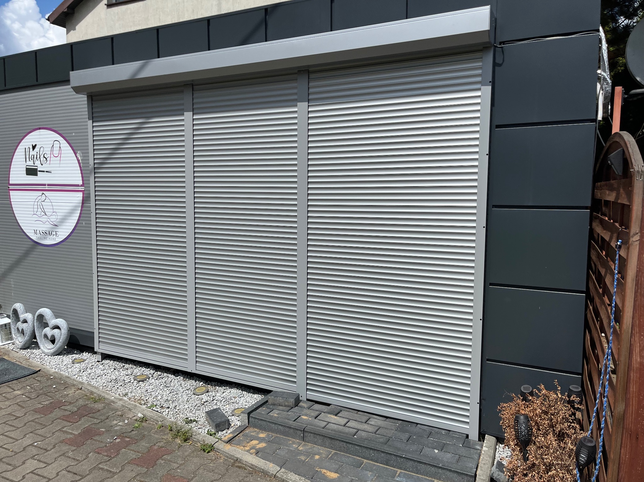 Zewnętrzne, srebrne rolety na oknach lokalu usługowego z szyldem 'Nails & Massage', dekoracyjne kamienne serca przed wejściem, widok z poziomu chodnika.
