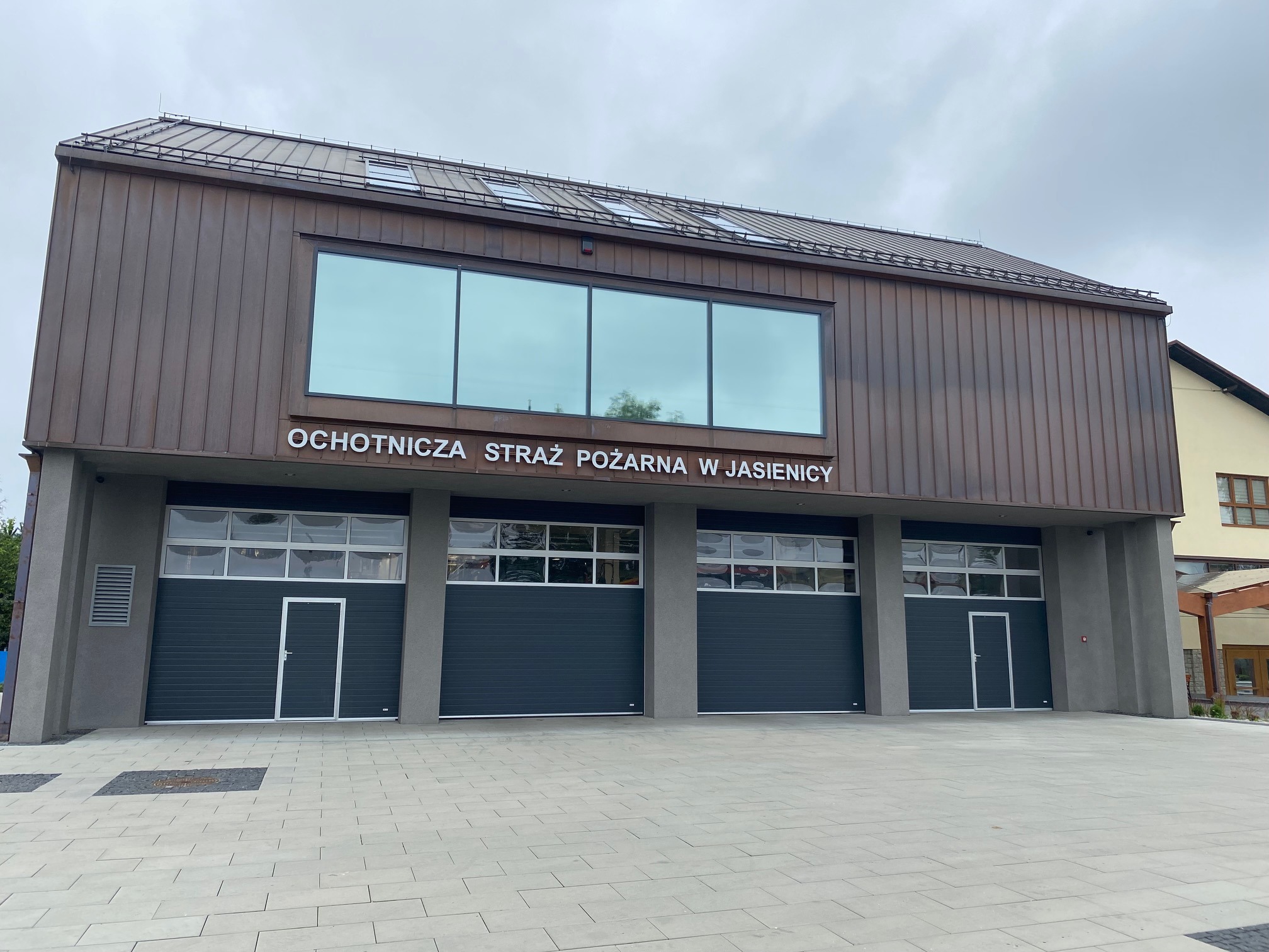 Nowoczesna remiza strażacka z dwoma segmentowymi bramami garażowymi w kolorze grafitowym, zintegrowanymi drzwiami wejściowymi i przeszkleniami w górnej części, elewacja w kolorze brązu.
