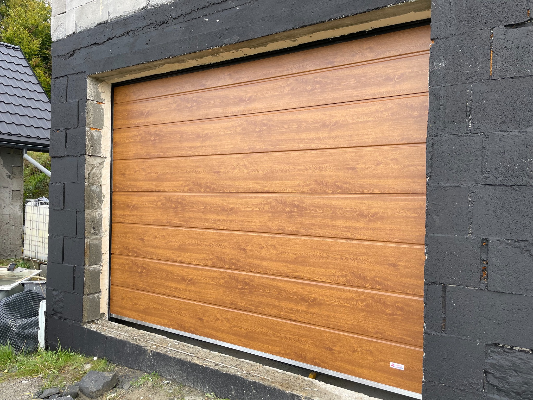 Zamknięta brama segmentowa garażowa w kolorze jasnego drewna, zamontowana w surowym otworze murowanym z pomalowanymi na czarno bloczkami betonowymi.