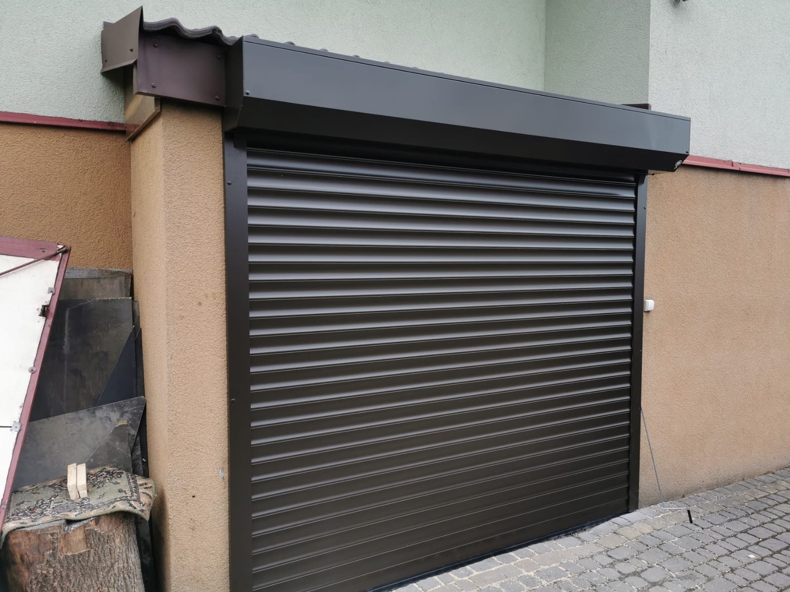 Zamknięta, brązowa brama rolowana garażowa z widocznymi elementami konstrukcyjnymi i fragmentem elewacji budynku.