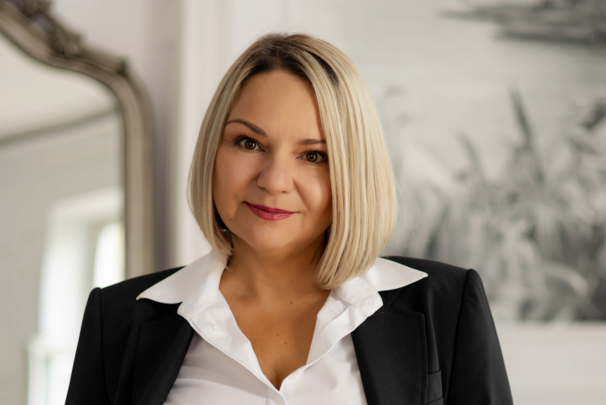 Portret kobiety w czarnej marynarce i białej koszuli, z blond włosami do ramion, patrzącej w obiektyw. W tle widoczne lustro i abstrakcyjny obraz.