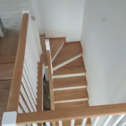 Widok z góry na jasne, dębowe schody z białymi podstopnicami i balustradą z białymi tralkami, w minimalistycznym wnętrzu z białymi ścianami.