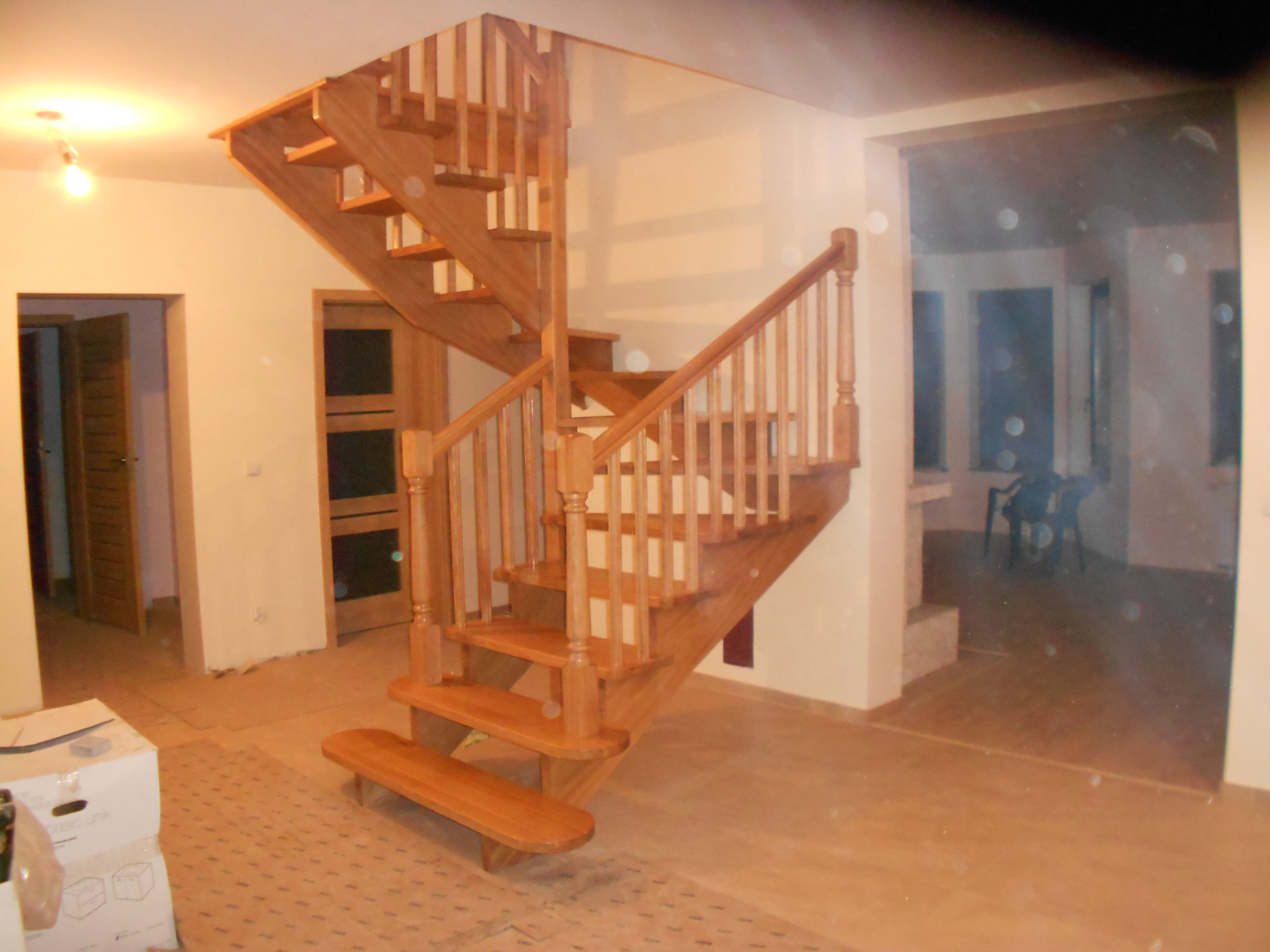 Jasne, drewniane schody spiralne z poręczą w trakcie montażu w nowym domu, widoczne kartony i niedokończone wykończenie podłogi.