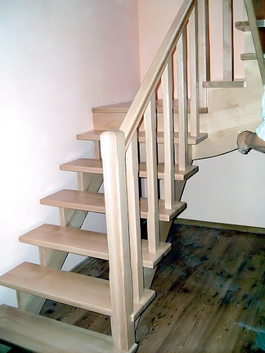 Jasne, drewniane schody z balustradą w trakcie montażu, widoczne niedoskonałości wykończenia i fragment dłoni pracownika.