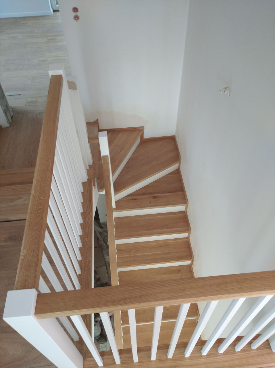 Widok z góry na jasne, dębowe schody z białymi podstopnicami i balustradą z białymi tralkami, w minimalistycznym wnętrzu z białymi ścianami.