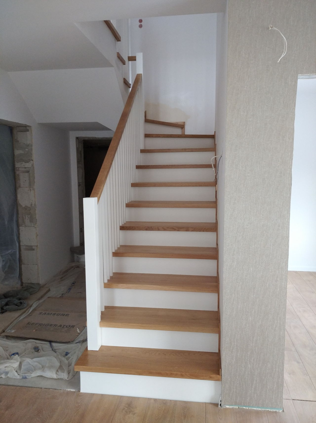 Nowe schody z jasnego drewna z białą balustradą, widoczne w trakcie montażu, z zabezpieczonym podłożem przed zabrudzeniem.
