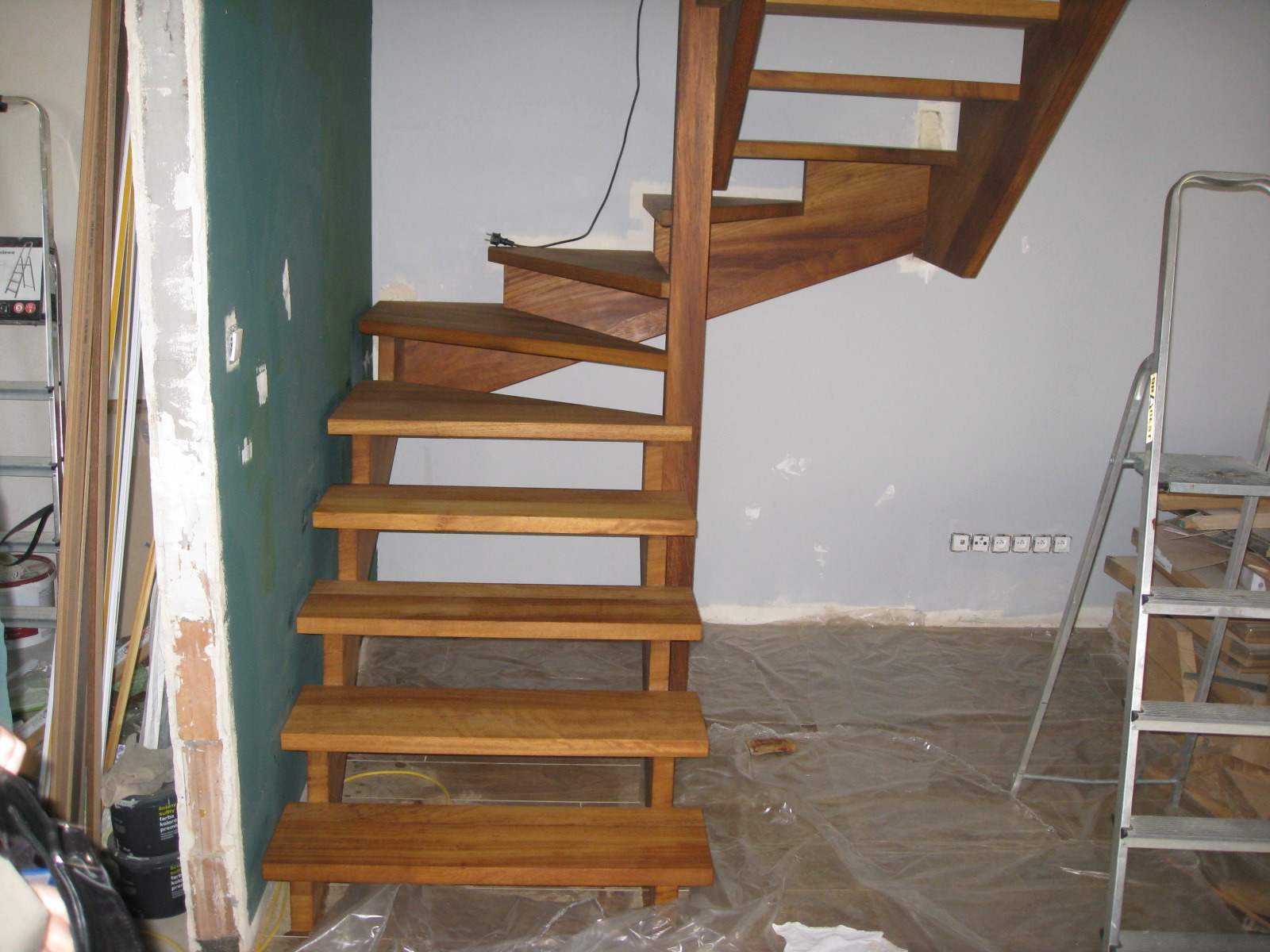 Wewnętrzne, spiralne stopnie z jasnego drewna podczas montażu, zabezpieczona folią podłoga, ściany w trakcie malowania, widoczna drabina i materiały budowlane.