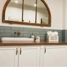 Umywalka nablatowa z baterią w kolorze mosiądzu, błękitne płytki ścienne, lustro w drewnianej ramie okiennej, białe szafki z podłużnymi uchwytami i dekoracyjne akcesoria łazienkowe.