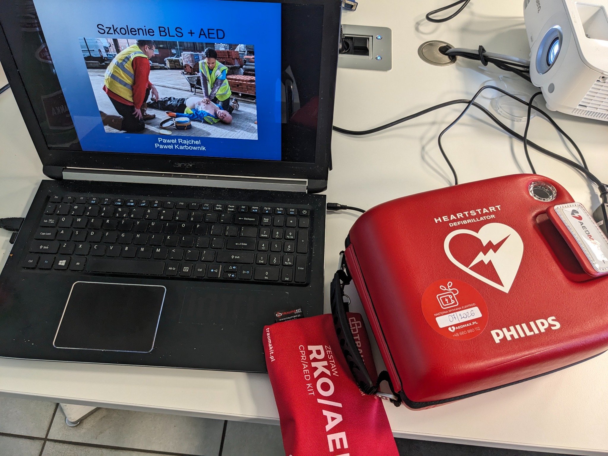 Laptop wyświetla prezentację ze szkolenia BLS+AED. Obok defibrylator Philips i zestaw RKO/AED. Widoczny projektor. Sprzęt na białym stole.