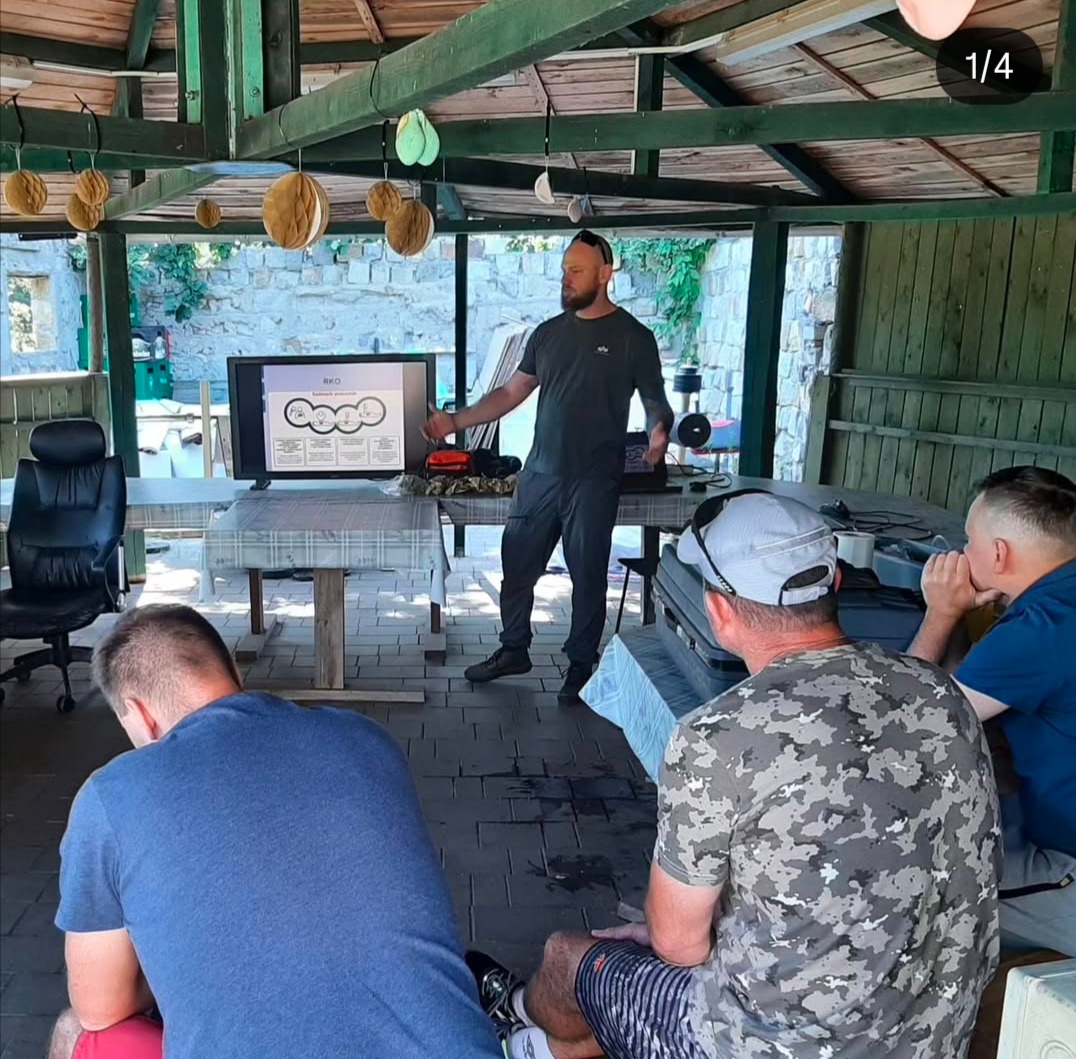 Instruktor prezentuje schemat RKO na monitorze podczas szkolenia w altanie. Uczestnicy siedzą przodem do ekranu, słuchając wykładu. Kamienna ściana w tle.