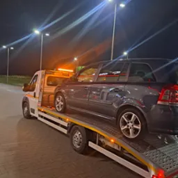 Ciemnoszary samochód osobowy marki Opel na platformie lawety, oświetlonej pomarańczowymi światłami ostrzegawczymi, w tle nocne oświetlenie uliczne.
