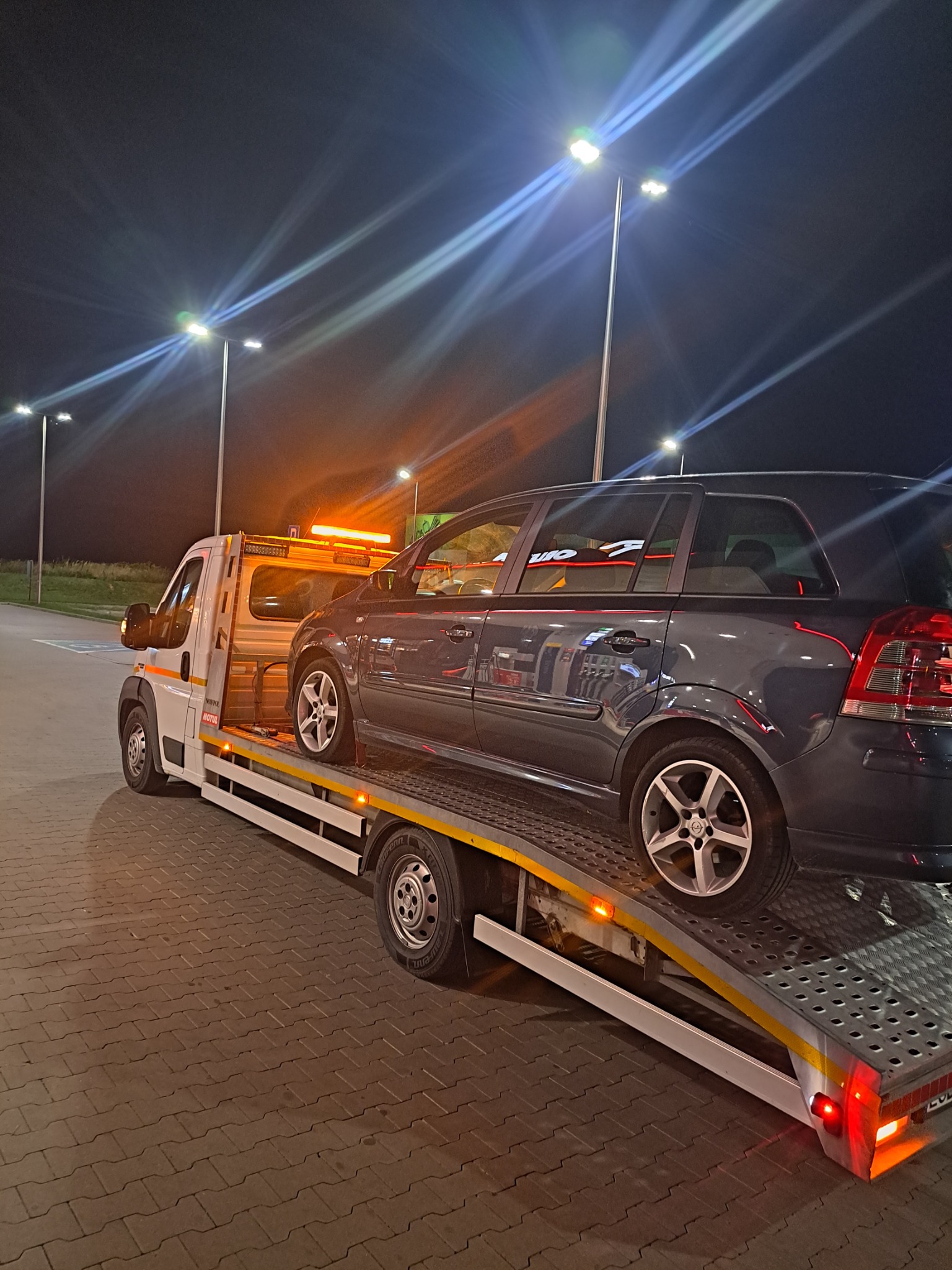 Ciemnoszary samochód osobowy marki Opel na platformie lawety, oświetlonej pomarańczowymi światłami ostrzegawczymi, w tle nocne oświetlenie uliczne.