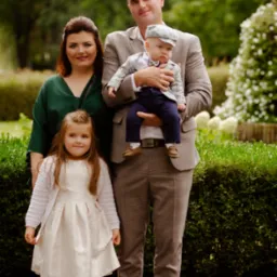 Rodzinne zdjęcie portretowe: ojciec w brązowym garniturze w kratę trzyma syna w podobnym stroju, obok matka w zielonej sukience i córka w jasnej sukience z bolerkiem, na tle zieleni.