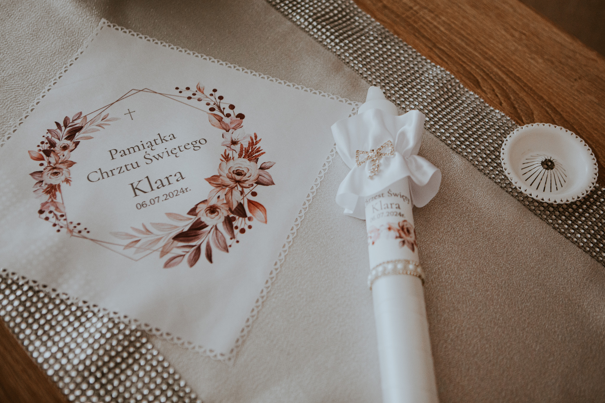 Pamiątka Chrztu Świętego Klary: biała serwetka z nadrukiem florystycznym i datą 06.07.2024, obok świeca z kokardką i imieniem Klara, ustawione na dekoracyjnym bieżniku.