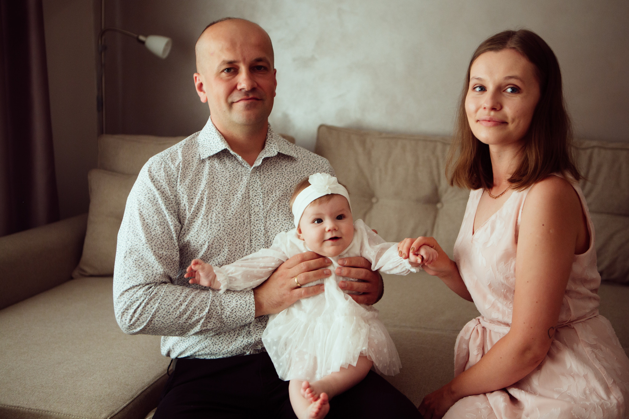 Rodzinne zdjęcie: ojciec trzyma na kolanach ubraną w białą sukienkę córeczkę, matka siedzi obok, trzymając dłoń dziecka, neutralne tło.