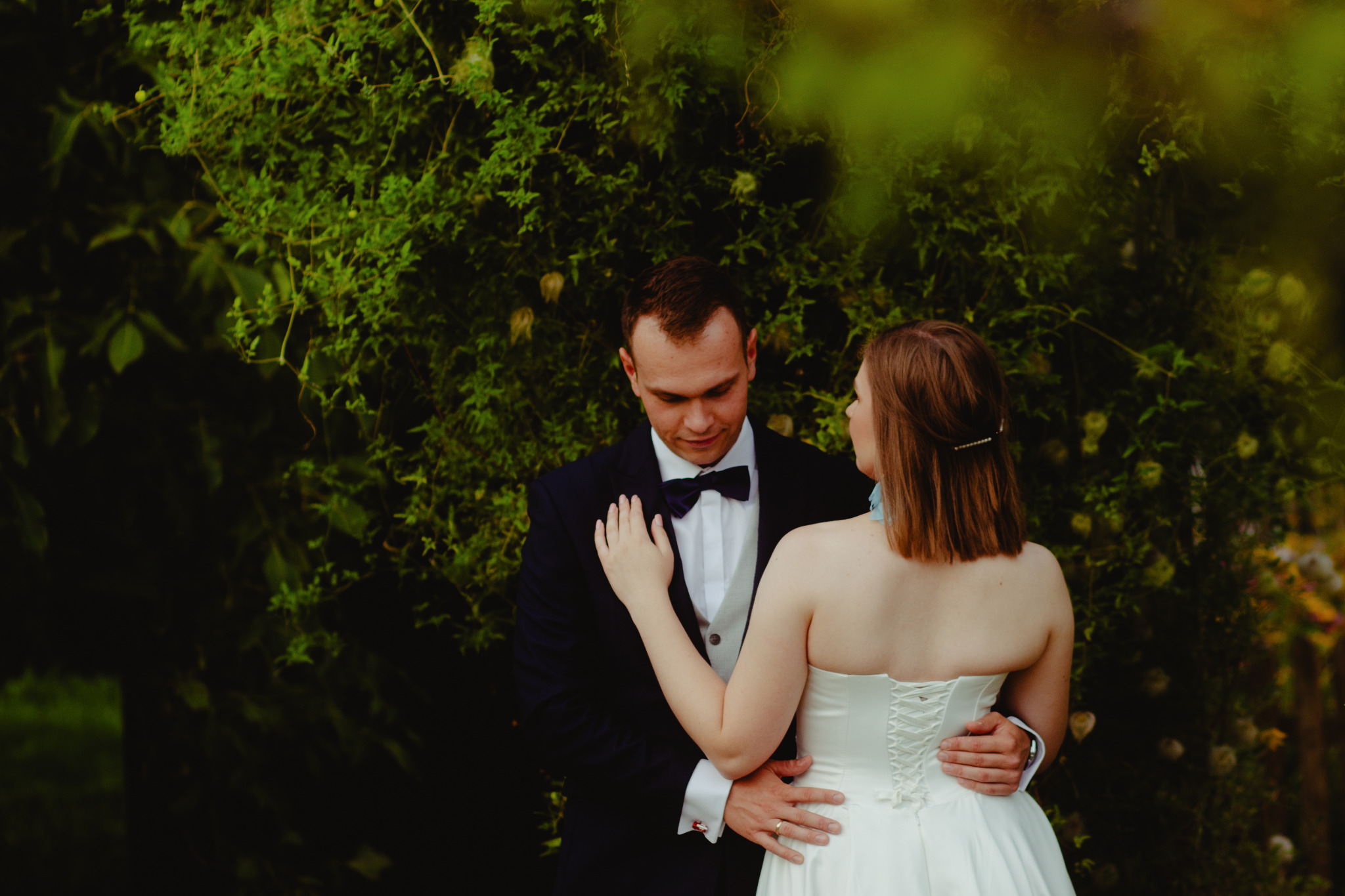 Para młoda w objęciach na tle zielonej ściany roślin, on w granatowym smokingu z muchą, ona w białej sukni z gorsetem, delikatne światło.