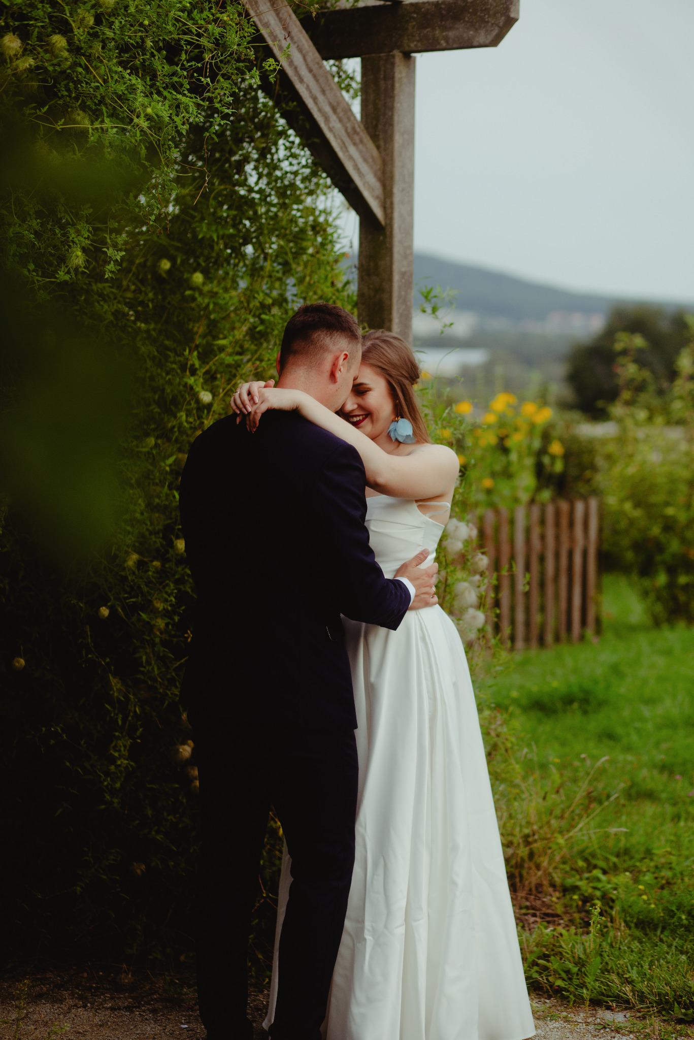 Para młoda obejmująca się pod drewnianą konstrukcją w ogrodzie, panna młoda w białej sukni z niebieskimi kolczykami, uśmiechnięta, w tle widok na wzgórza i żółte kwiaty.