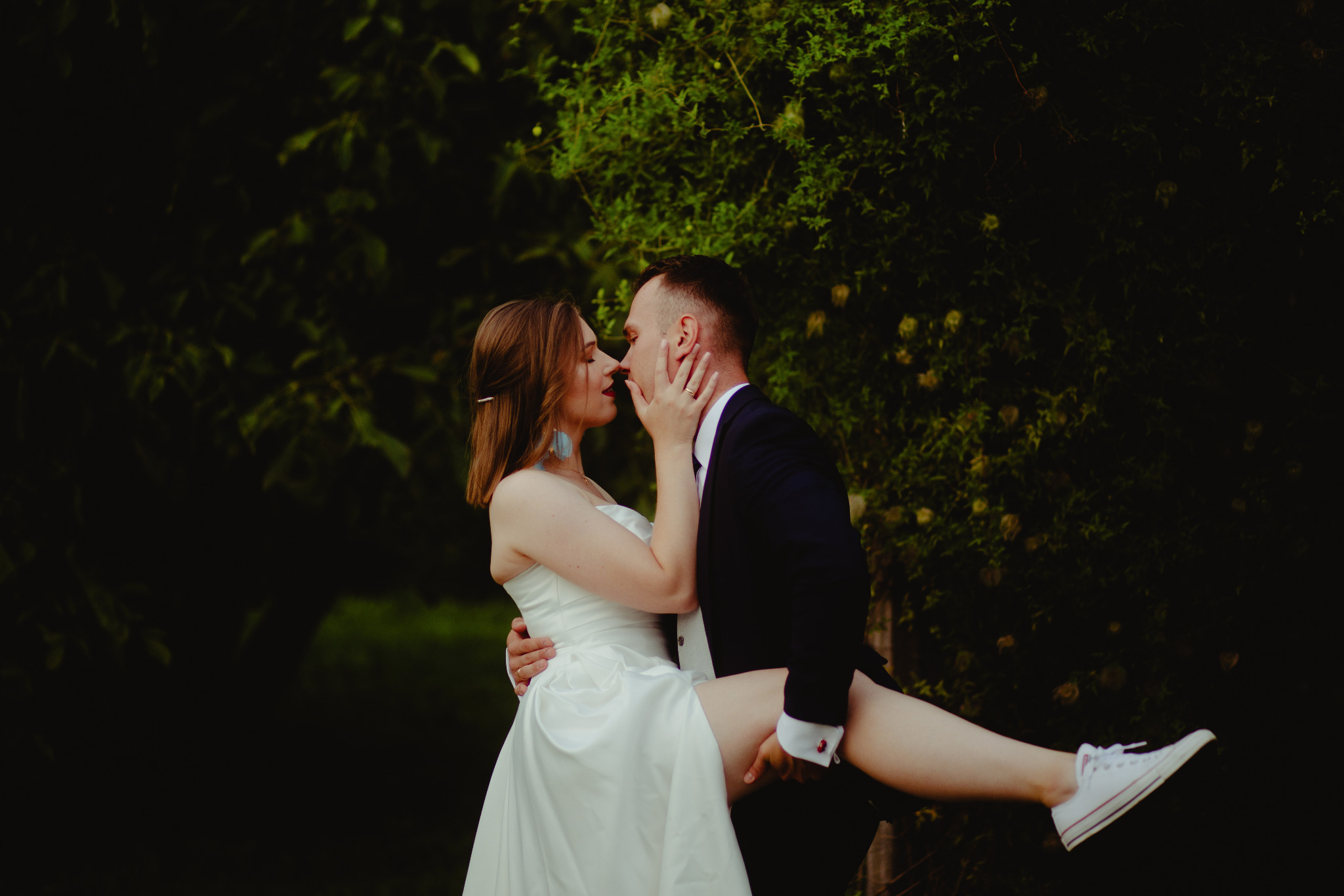 Pan młody trzyma pannę młodą w białej sukni i białych trampkach na tle zieleni, zbliżają się do pocałunku.