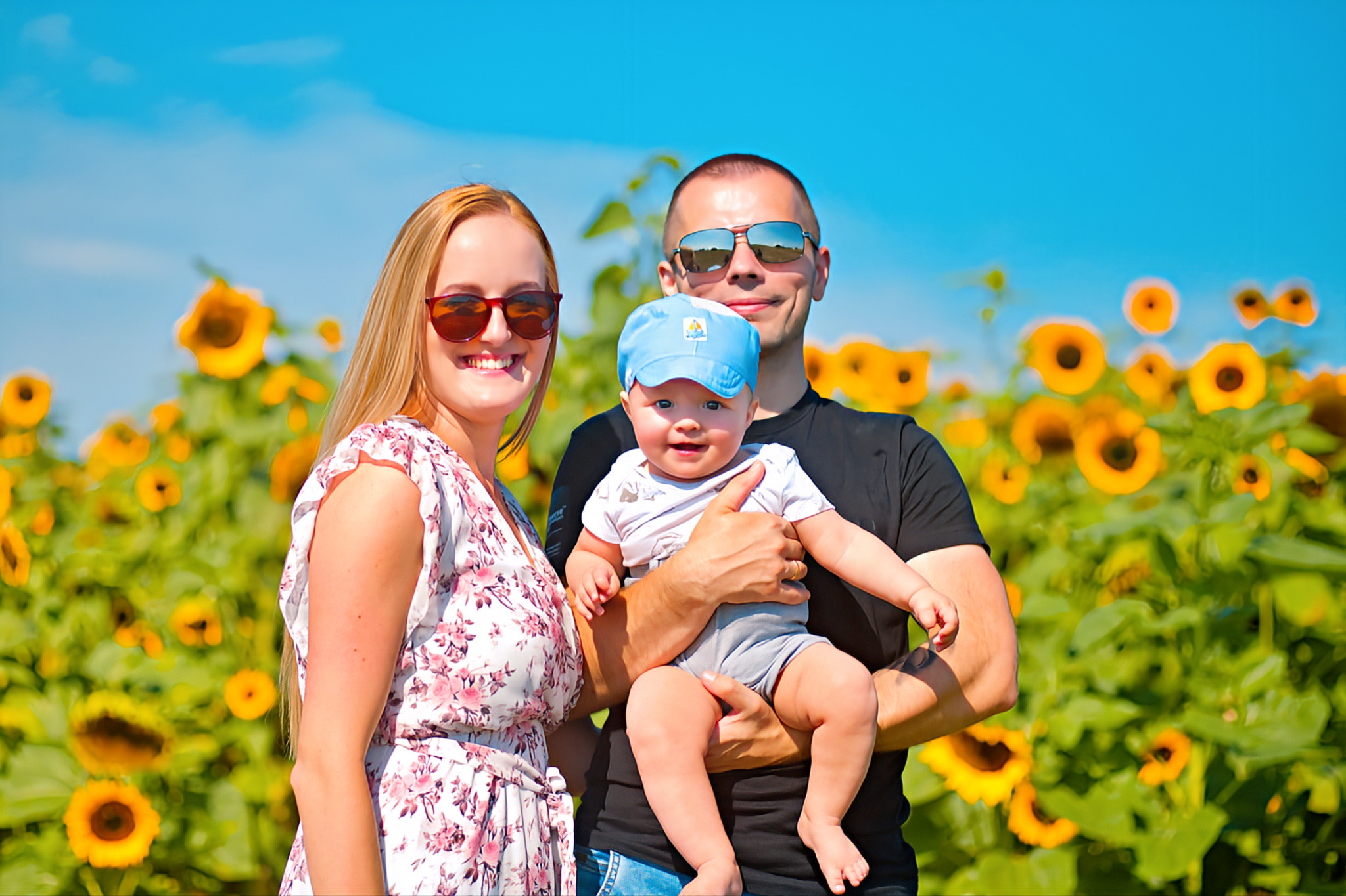 Uśmiechnięta rodzina: mama w kwiecistej sukience, tata w czarnej koszulce trzymający na rękach roześmianego synka w niebieskiej czapce, na tle pola słoneczników i błękitnego nieba.