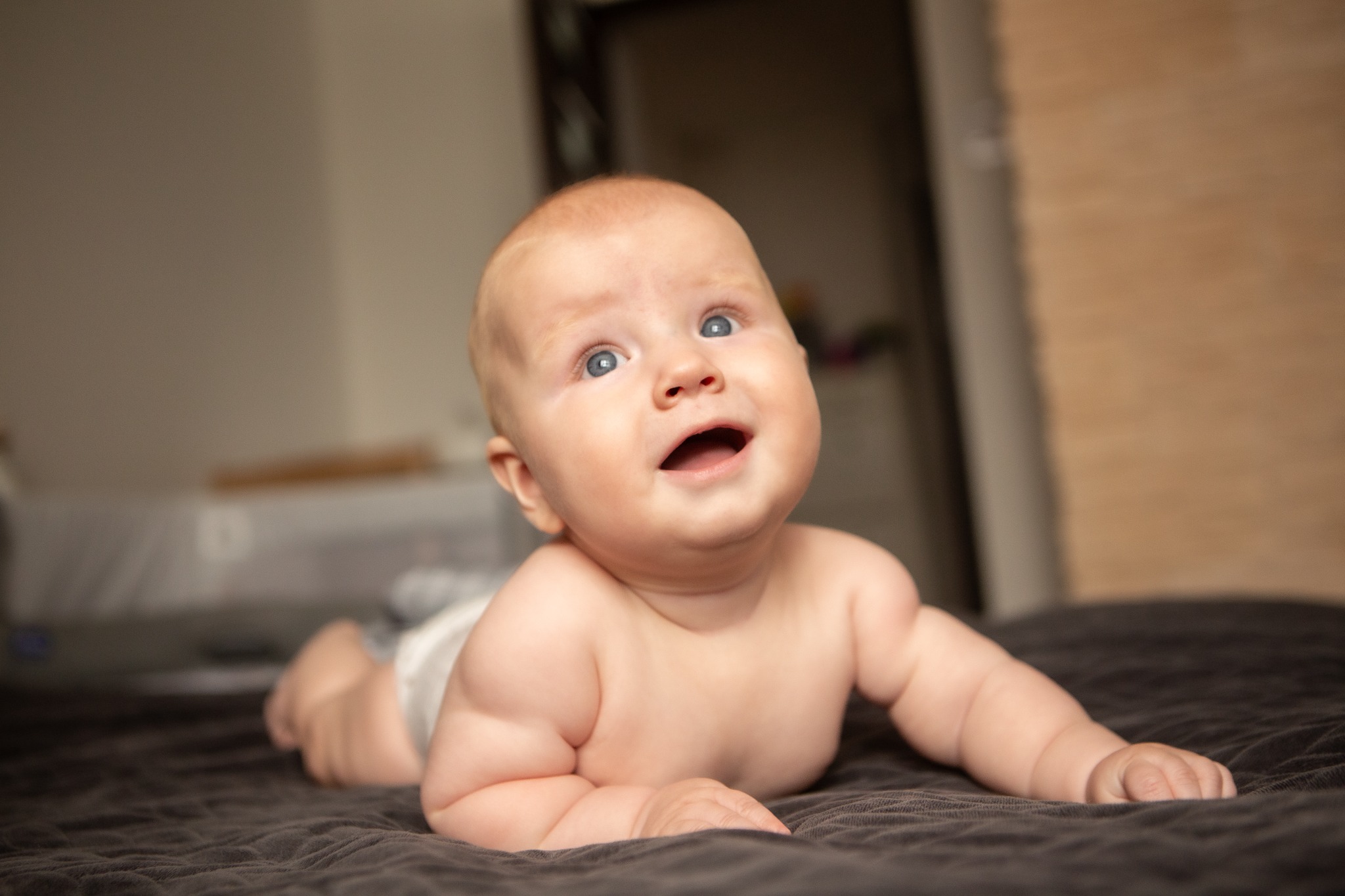 Niemowlę leżące na brzuchu na ciemnoszarym, teksturowanym kocu, z zadartą głową i otwartymi ustami, patrzące w górę, z widocznymi niebieskimi oczami i bladą skórą.