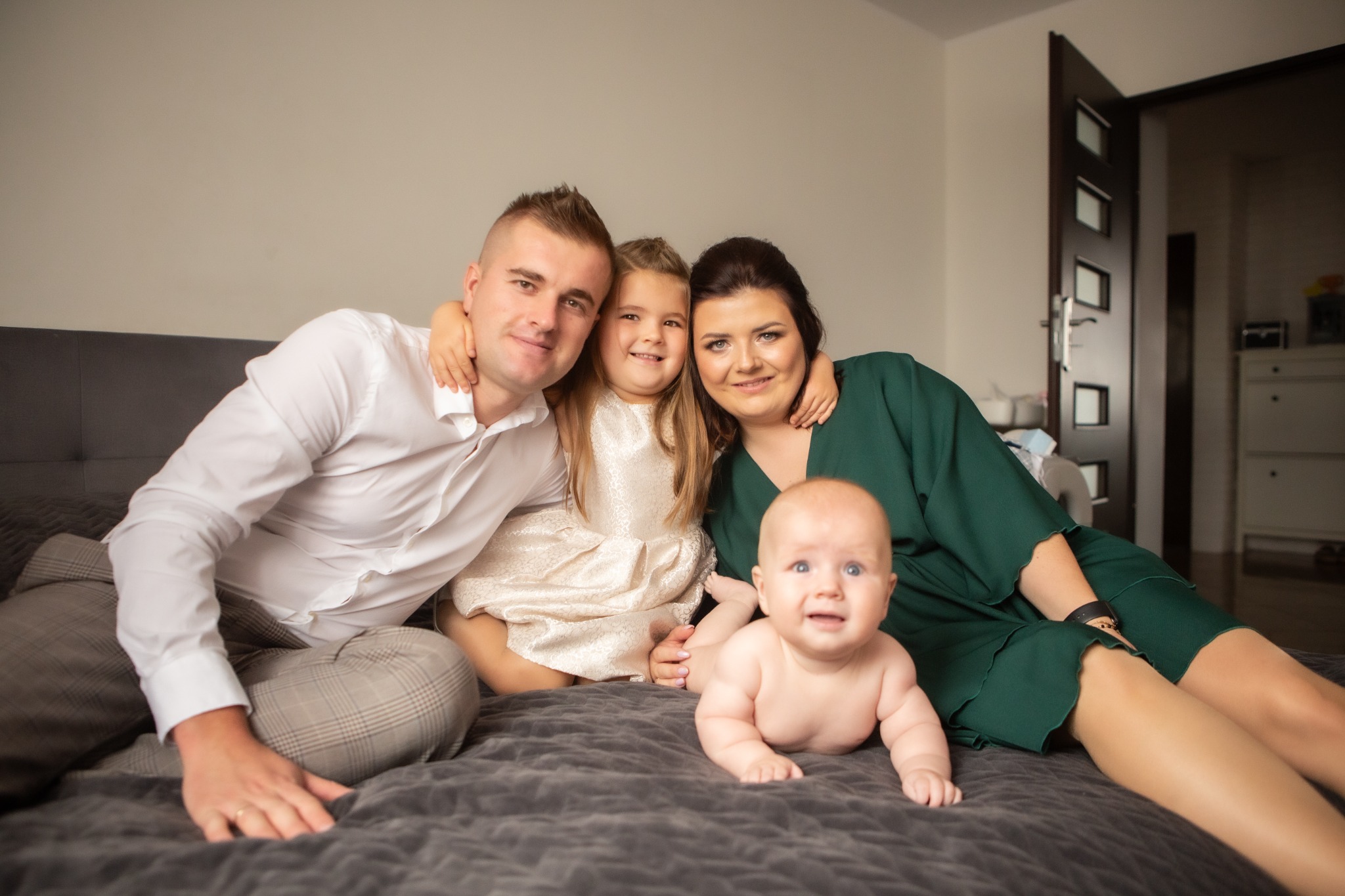 Rodzinne zdjęcie na łóżku: tata w białej koszuli i kraciastych spodniach, mama w zielonej sukience, córka w kremowej sukience z koronką i niemowlę leżące na brzuchu, patrzące w obiektyw.