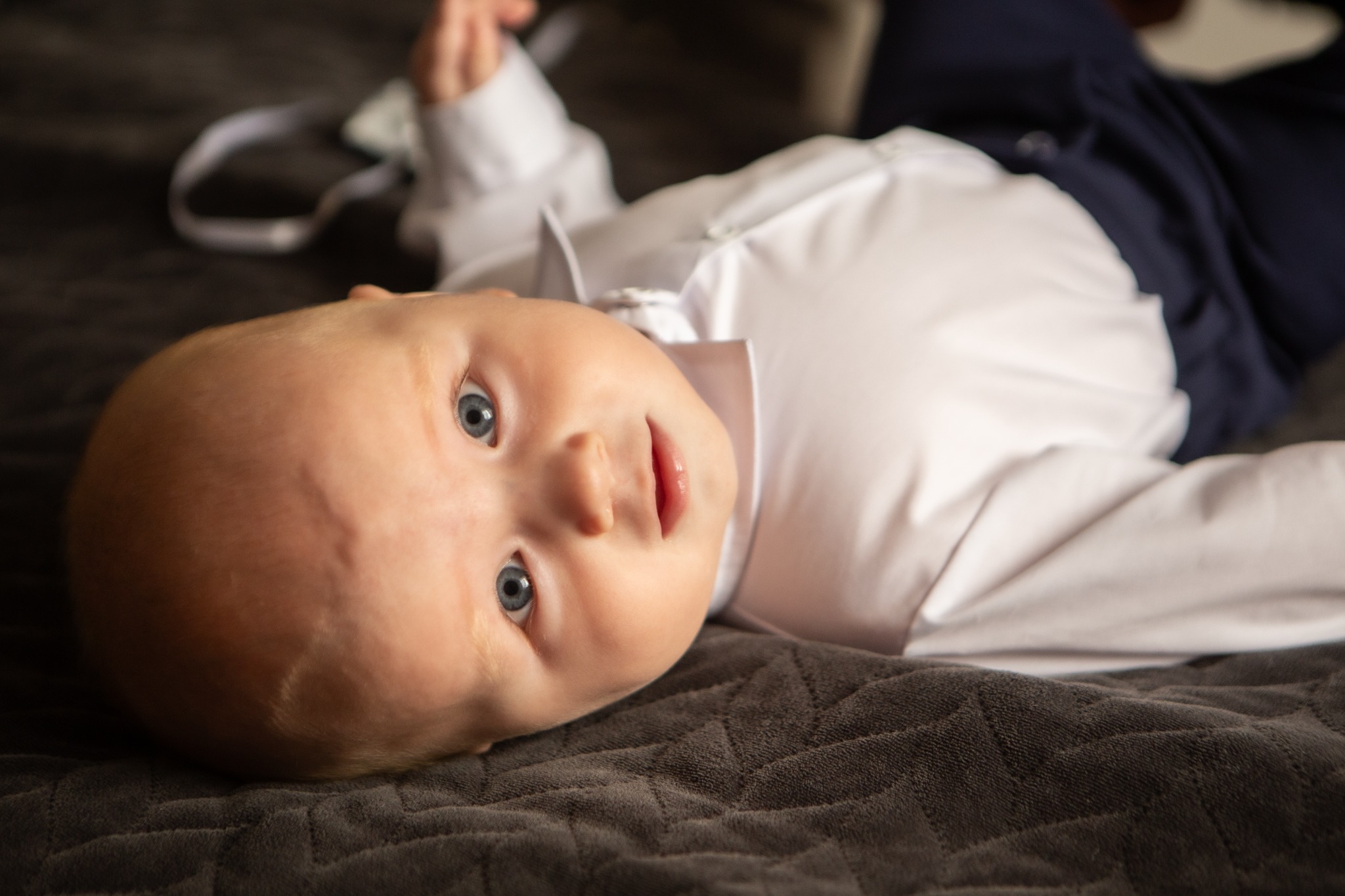 Niemowlę w białej koszuli i granatowych spodniach leży na szarym kocu, patrząc w górę, zdjęcie wykonane w Skarżysko Kościelne.