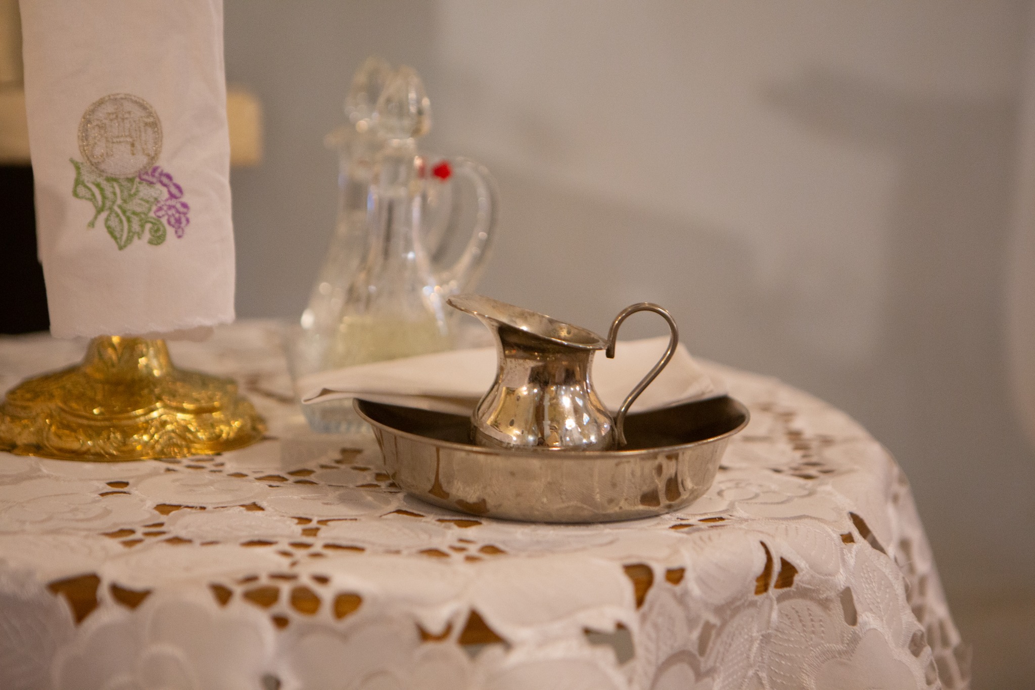 Srebrny dzbanuszek na wodę święconą w miseczce, złota podstawa pod białą serwetą z haftem eucharystycznym, na białym obrusie z ażurowym wzorem.