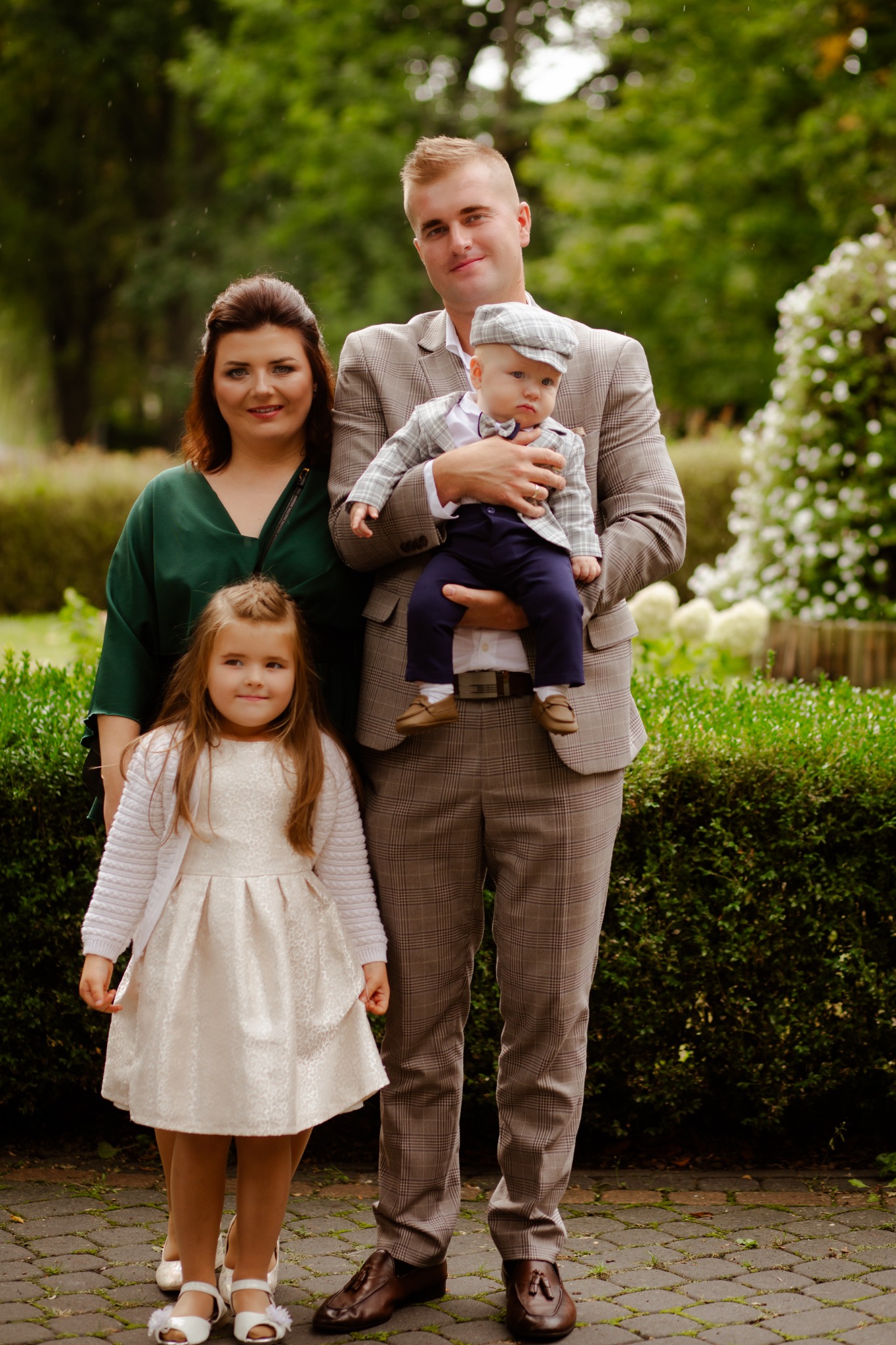 Rodzinne zdjęcie portretowe: ojciec w brązowym garniturze w kratę trzyma syna w podobnym stroju, obok matka w zielonej sukience i córka w jasnej sukience z bolerkiem, na tle zieleni.