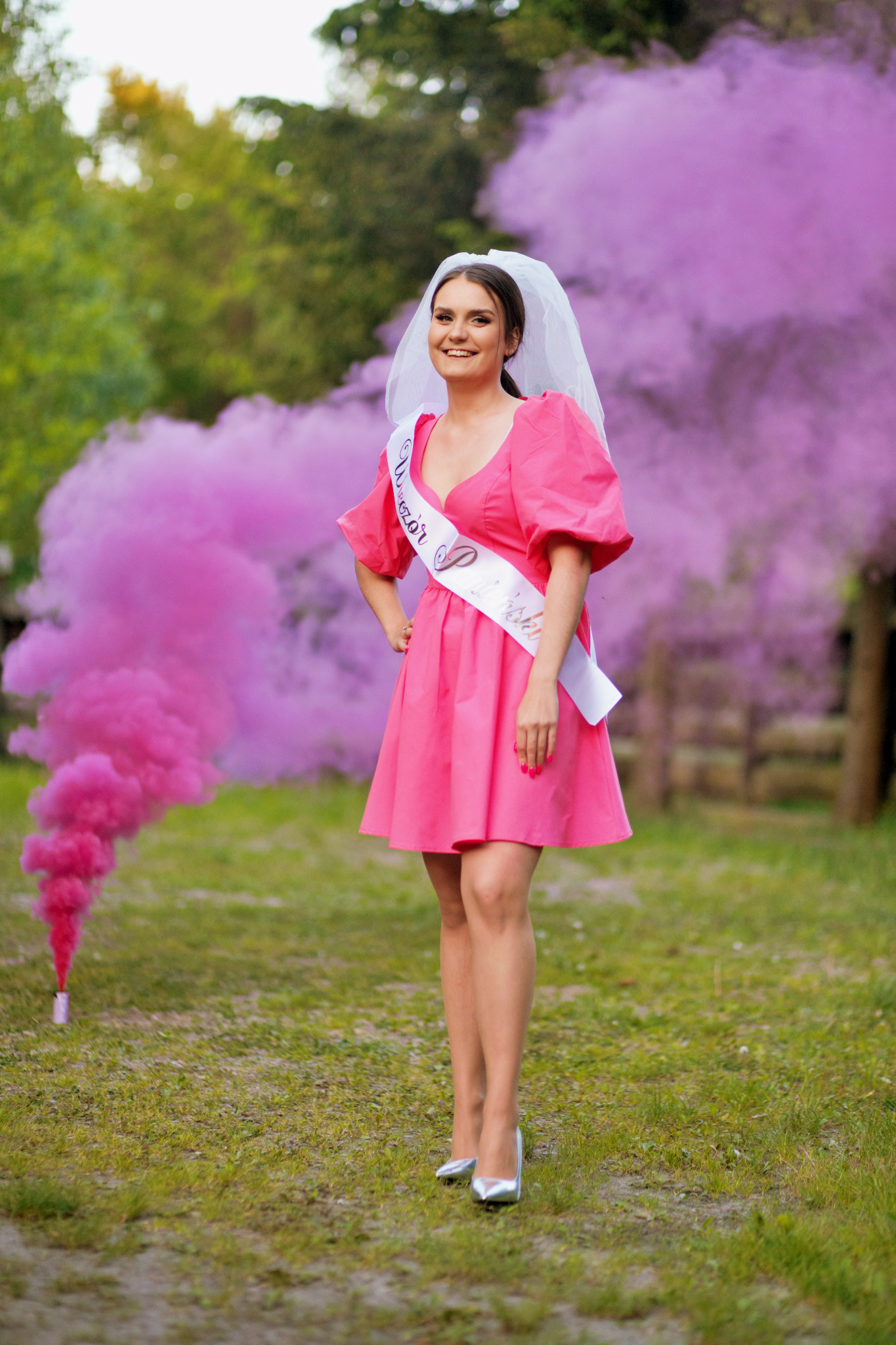 Uśmiechnięta kobieta w różowej sukience, welonie i szarfie 'Wieczór Panieński' pozuje na łące w Skarżysku Kościelnym, otoczona obłokami różowego dymu.