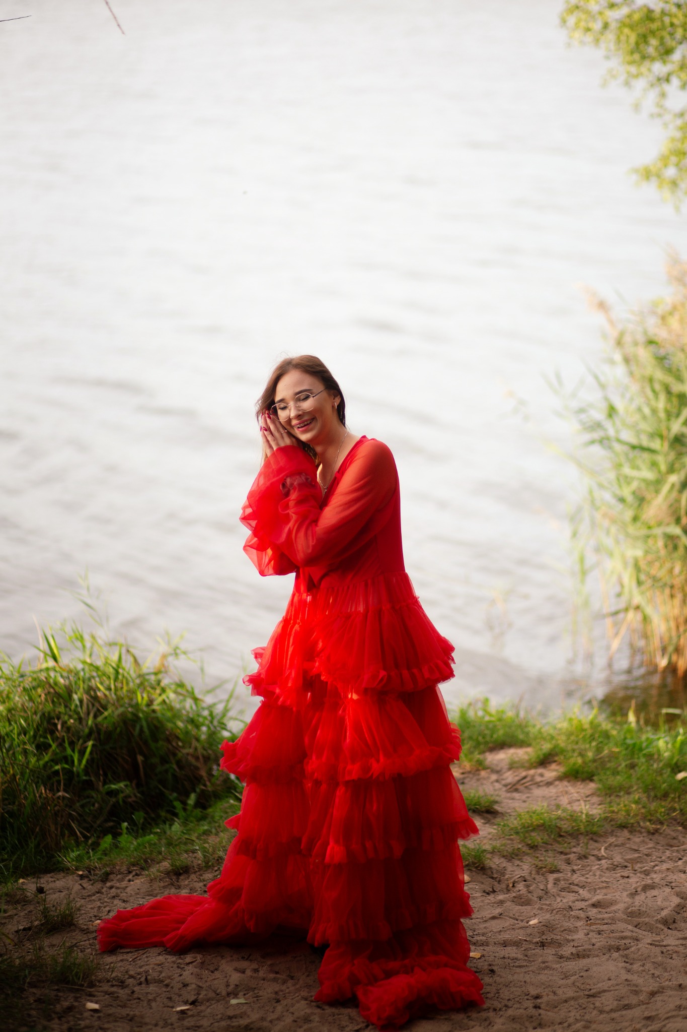 Uśmiechnięta kobieta w okularach i czerwonej, tiulowej sukni pozuje na brzegu jeziora.