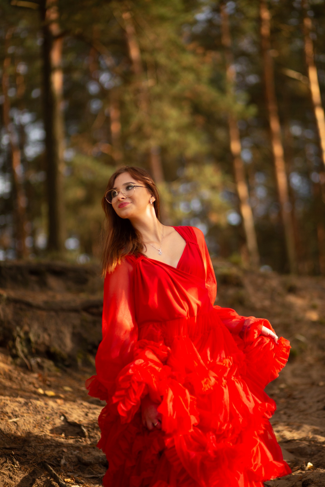 Kobieta w zwiewnej, czerwonej sukni z falbanami, okularach i srebrnym naszyjniku pozuje na tle leśnej ścieżki w promieniach zachodzącego słońca.