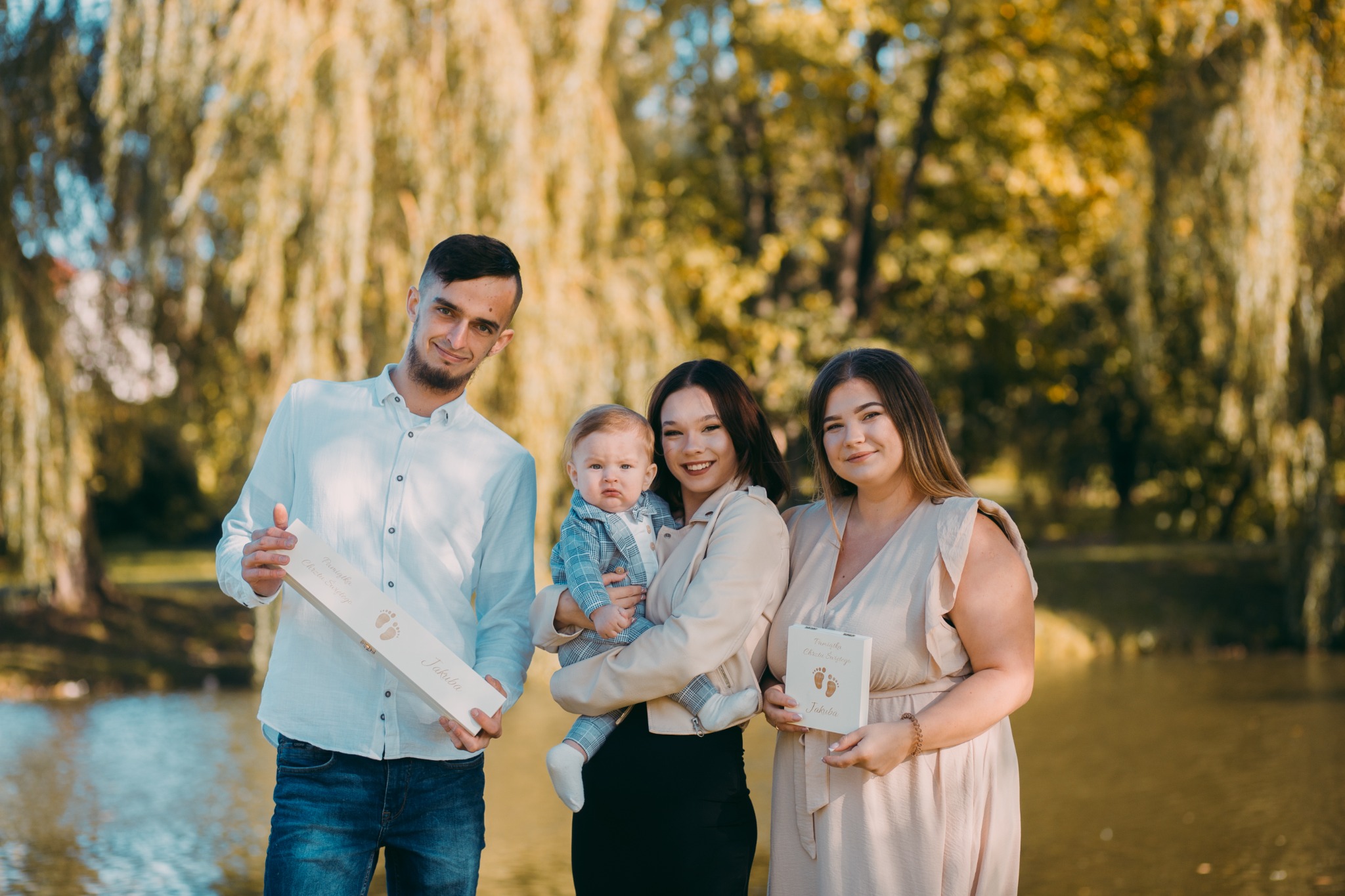 Uśmiechnięta rodzina z małym dzieckiem pozuje nad wodą, trzymając pamiątkowe pudełka z nadrukiem stóp dziecka i napisem 'Pamiątka Chrztu Świętego Jakuba'.