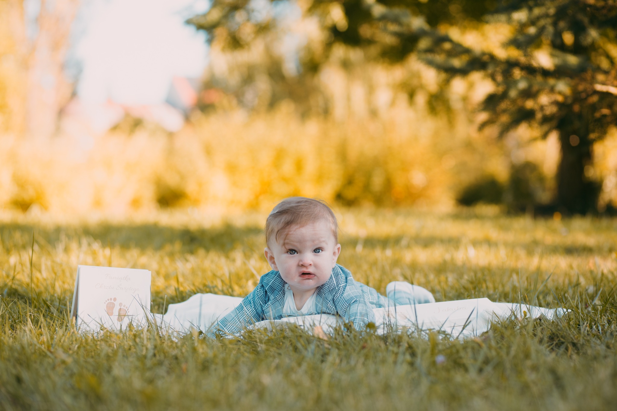 Niemowlę w niebieskiej marynarce leży na białym kocyku na trawie obok pamiątki Chrztu Świętego z odciśniętymi stópkami, na tle zamazanych drzew.