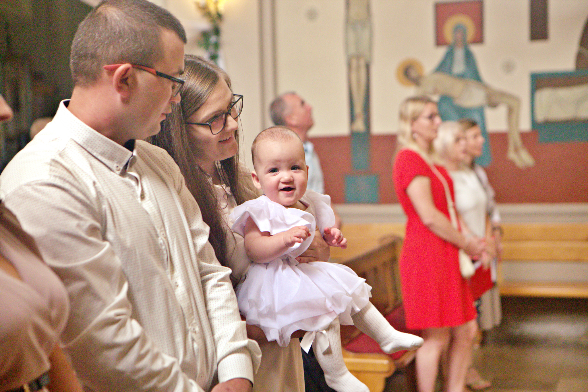 Uśmiechnięta dziewczynka w białej sukience i podkolanówkach, trzymana na rękach przez kobietę w kościele podczas ceremonii, z widocznymi gośćmi i obrazem religijnym w tle.
